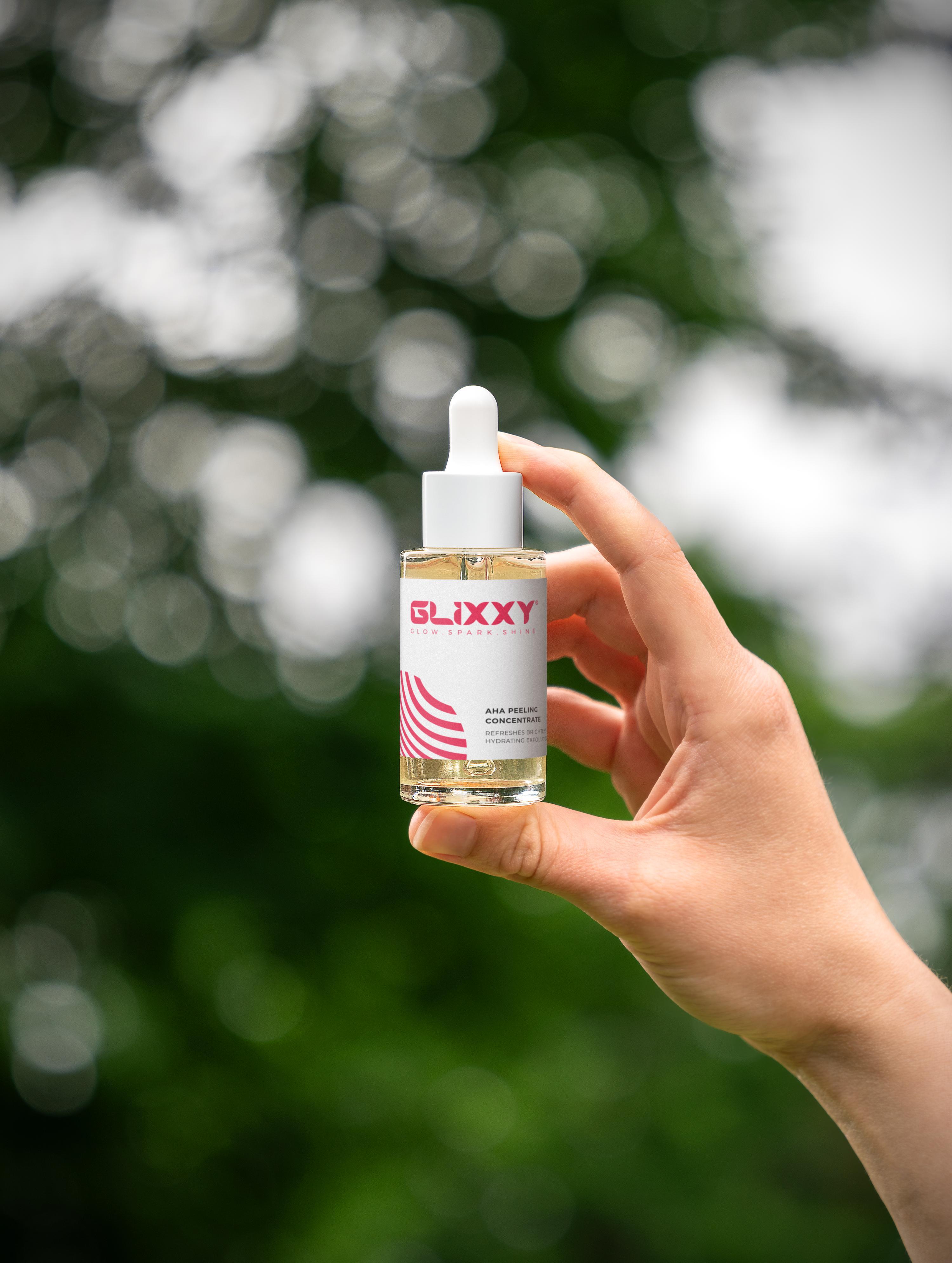 Hand holding a Glixxy aha peeling concentrate dropper bottle against a blurred green background