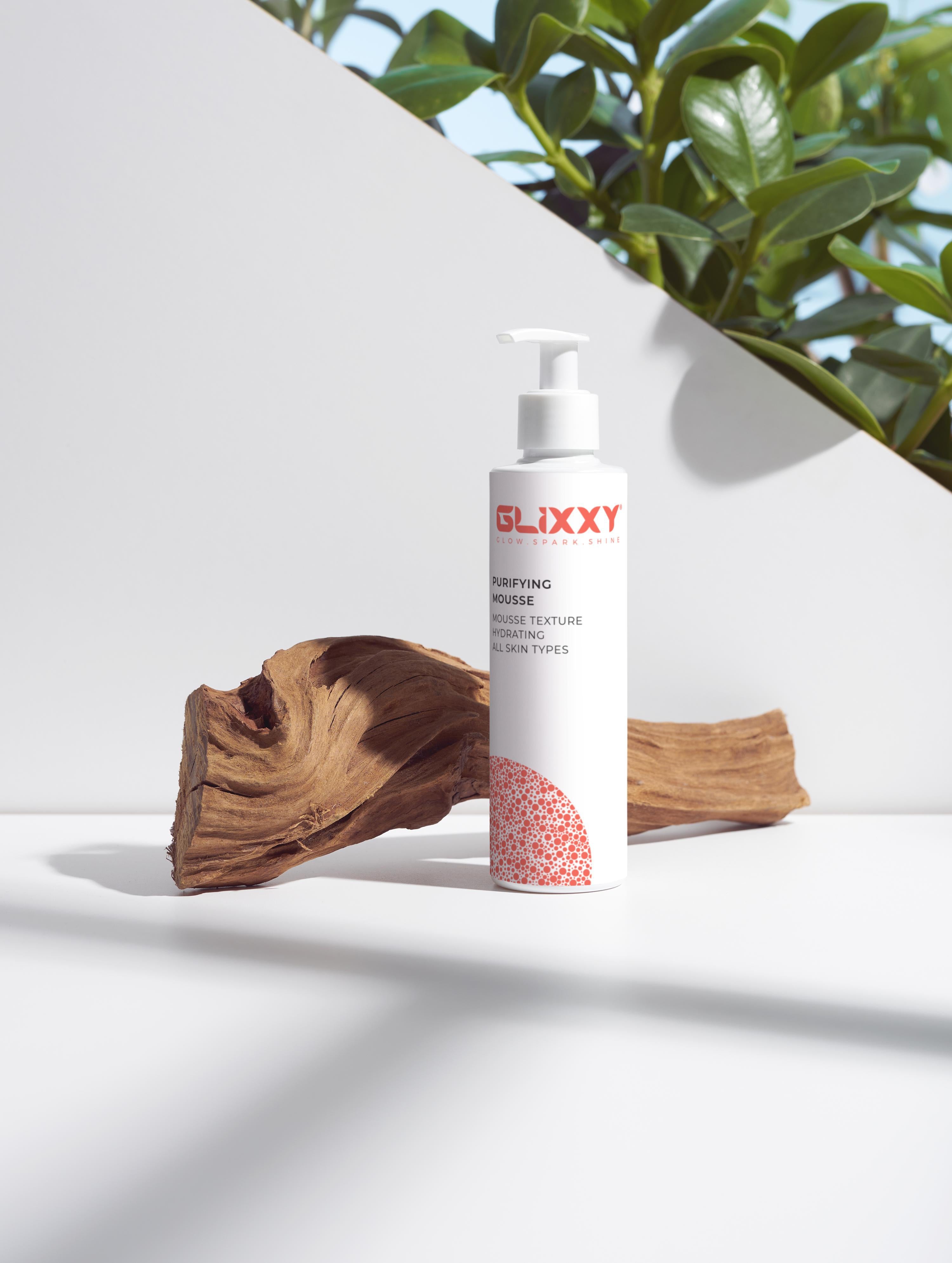 Glixxy purifying mousse bottle with pink cap and label on a white surface with natural elements