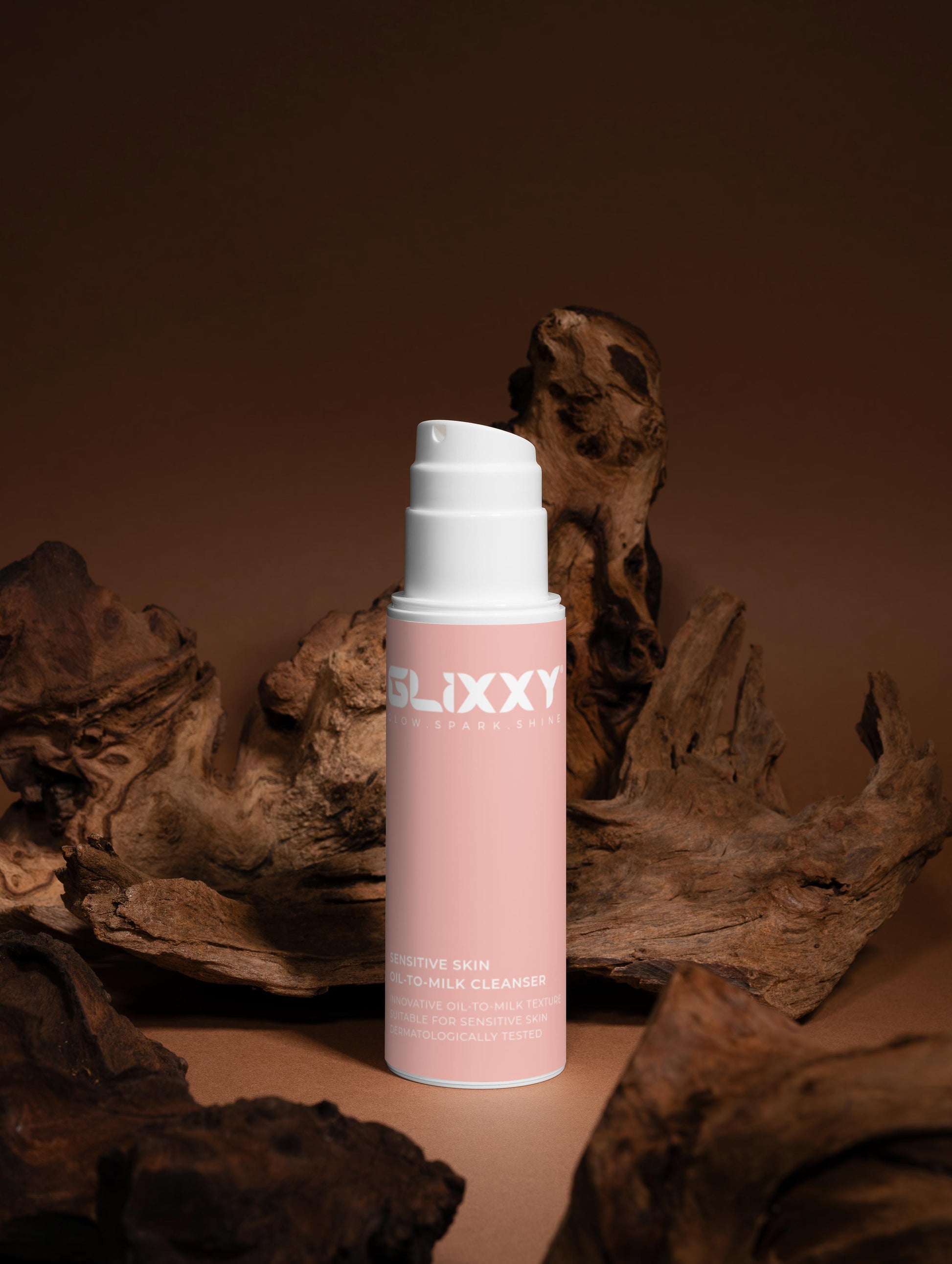 Pink Sensitive Skin Oil-To-Milk Cleanser bottle with white cap labeled 'Glixxy' against a brown background with wooden elements.